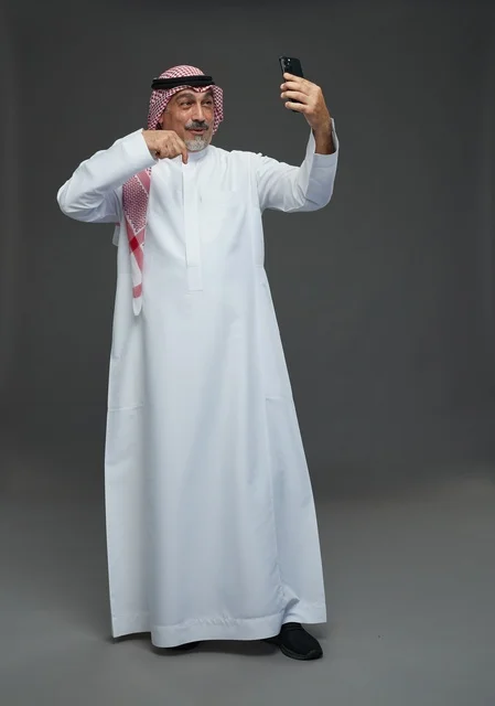 رجل  عربي سعودي خليجي مسن يرتدي الزي الثوب الابيض والشماغ ويمسك الهاتف المحمول, التقاط صورة سيلفي, اجراء مكالمة فيديو عبر الجوال,  ايماءات واشارات مختلفه , كامل الجسم, خلفيه رماديه