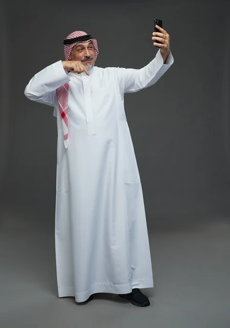 An elderly Arab Saudi Gulf man wearing a white thobe and a shemagh, holding a mobile phone, taking a selfie, making a video call via the phone, with various gestures and signals, full body, gray background.