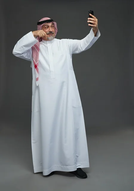 An elderly Arab Saudi Gulf man wearing a white thobe and a shemagh, holding a mobile phone, taking a selfie, making a video call via the phone, with various gestures and signals, full body, gray background.