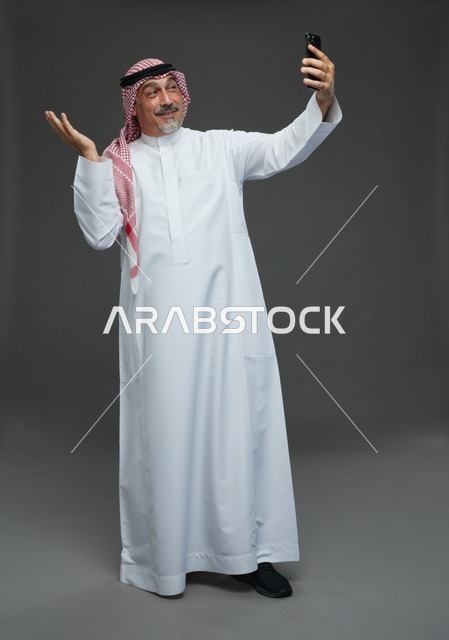 رجل  عربي سعودي خليجي مسن يرتدي الزي الثوب الابيض والشماغ ويمسك الهاتف المحمول, التقاط صورة سيلفي, اجراء مكالمة فيديو عبر الجوال,  ايماءات واشارات مختلفه , كامل الجسم, خلفيه رماديه