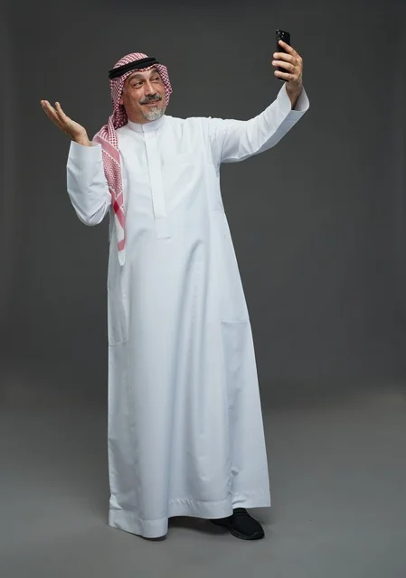 An elderly Arab Saudi Gulf man wearing a white thobe and a shemagh, holding a mobile phone, taking a selfie, making a video call via the phone, with various gestures and signals, full body, gray background.