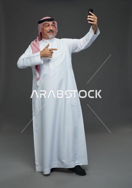An elderly Arab Saudi Gulf man wearing a white thobe and a shemagh, holding a mobile phone, taking a selfie, making a video call via the phone, with various gestures and signals, full body, gray background.