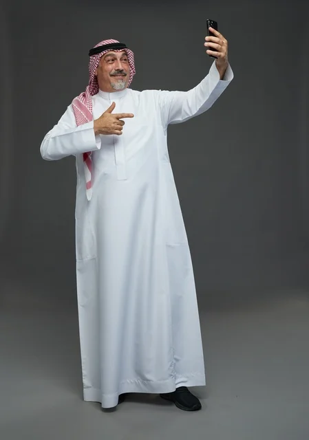 An elderly Arab Saudi Gulf man wearing a white thobe and a shemagh, holding a mobile phone, taking a selfie, making a video call via the phone, with various gestures and signals, full body, gray background.