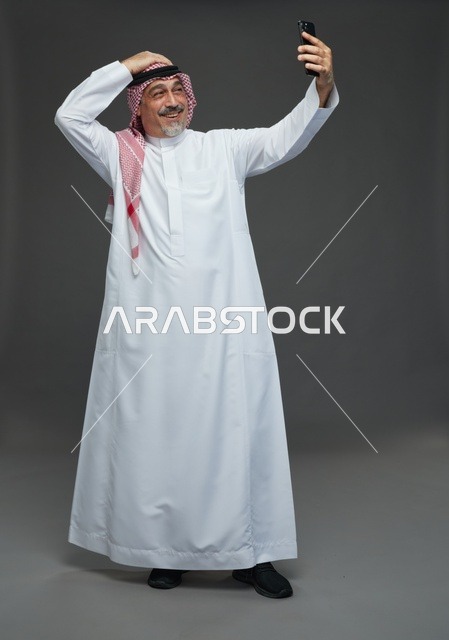An elderly Arab Saudi Gulf man wearing a white thobe and a shemagh, holding a mobile phone, taking a selfie, making a video call via the phone, with various gestures and signals, full body, gray background.