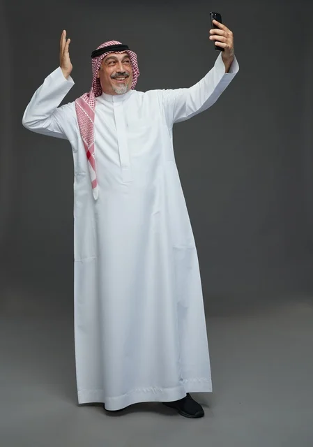 رجل  عربي سعودي خليجي مسن يرتدي الزي الثوب الابيض والشماغ ويمسك الهاتف المحمول, التقاط صورة سيلفي, اجراء مكالمة فيديو عبر الجوال,  ايماءات واشارات مختلفه , كامل الجسم, خلفيه رماديه