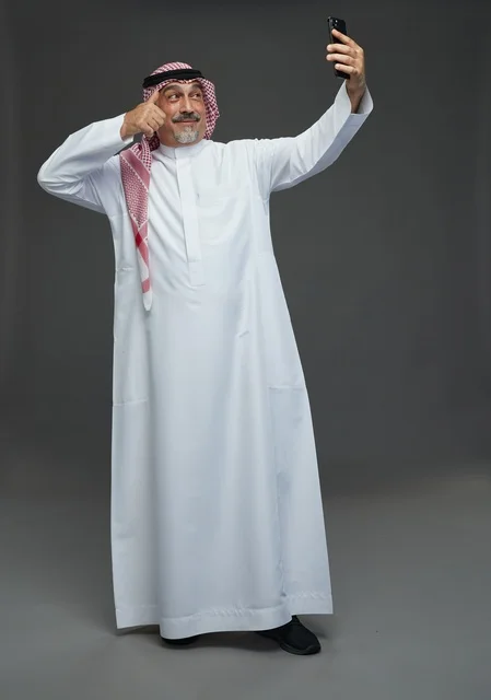 An elderly Arab Saudi Gulf man wearing a white thobe and a shemagh, holding a mobile phone, taking a selfie, making a video call via the phone, with various gestures and signals, full body, gray background.