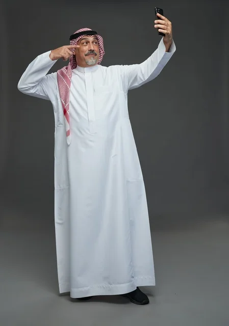 An elderly Arab Saudi Gulf man wearing a white thobe and a shemagh, holding a mobile phone, taking a selfie, making a video call via the phone, with various gestures and signals, full body, gray background.