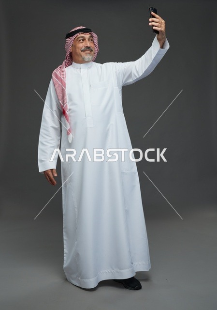 رجل  عربي سعودي خليجي مسن يرتدي الزي الثوب الابيض والشماغ ويمسك الهاتف المحمول, التقاط صورة سيلفي, اجراء مكالمة فيديو عبر الجوال,  ايماءات واشارات مختلفه , كامل الجسم, خلفيه رماديه