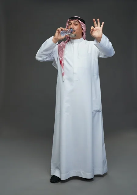 An elderly Arab Saudi Gulf man wearing a white thobe and a shemagh, holding a water bottle, with various gestures and signs, full body, gray background.