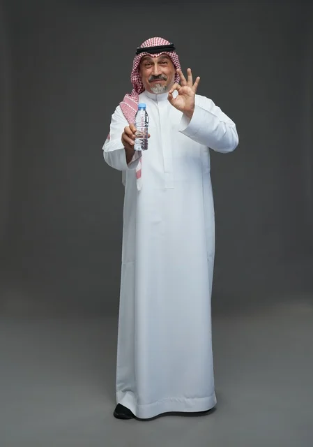 رجل  عربي سعودي خليجي مسن يرتدي الزي الثوب الابيض والشماغ ويحمل قارورة ماء,  ايماءات واشارات مختلفه , كامل الجسم, خلفيه رماديه