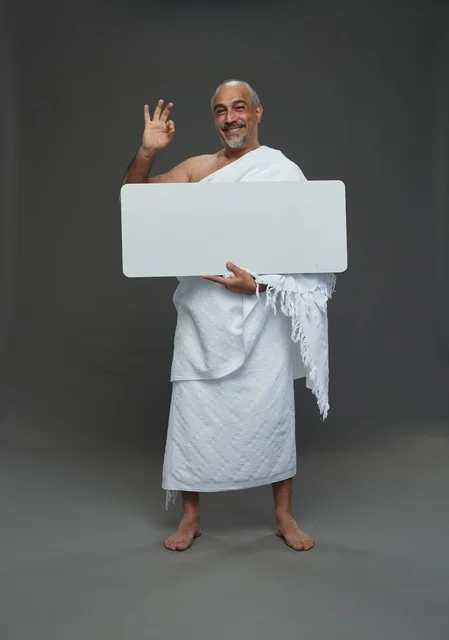 بورتريه لحاج سعودي بملابس الإحرام يحمل لوح مربع ابيض فارغ أثناء أداء المناسك, ايماءات واشارات مختلفه, كامل الجسم, خلفيه رماديه