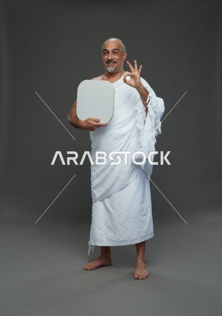 A portrait of a Saudi pilgrim in Ihram clothing holding a blank white square board while performing the rituals, with various gestures and signs, full body, gray background.