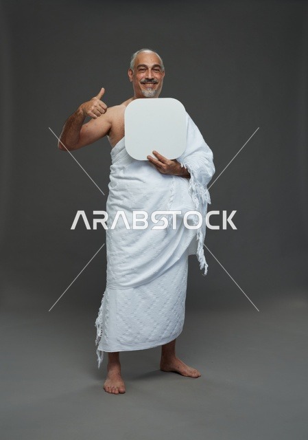 بورتريه لحاج سعودي بملابس الإحرام يحمل لوح مربع ابيض فارغ أثناء أداء المناسك, ايماءات واشارات مختلفه, كامل الجسم, خلفيه رماديه