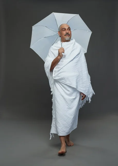 A portrait of a Saudi man in Ihram clothing holding a white umbrella while performing rituals, with various gestures and signals, full body, gray background.