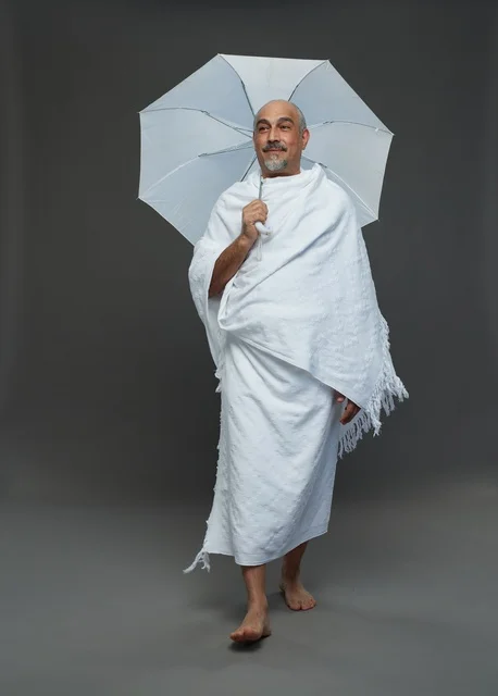 A portrait of a Saudi man in Ihram clothing holding a white umbrella while performing rituals, with various gestures and signals, full body, gray background.