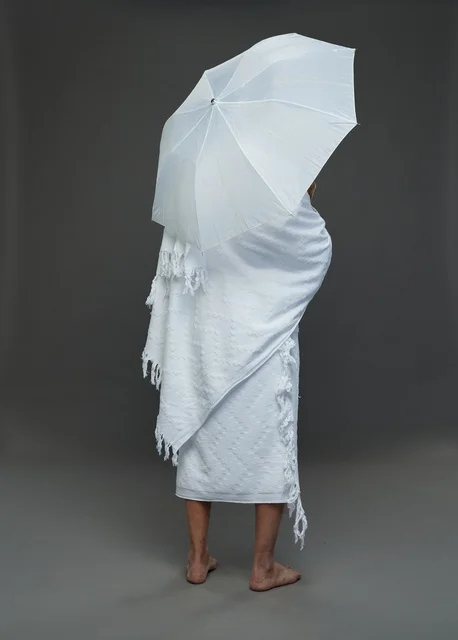 بورتريه لرجل سعودي بملابس الإحرام يحمل مظلة بيضاء أثناء أداء المناسك, ايماءات واشارات مختلفه, كامل الجسم, خلفيه رماديه