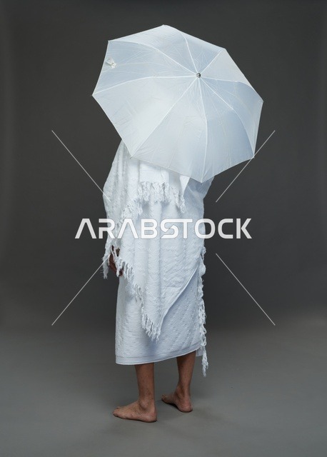 A portrait of a Saudi man in Ihram clothing holding a white umbrella while performing rituals, with various gestures and signals, full body, gray background.