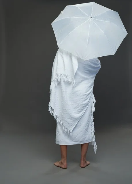 بورتريه لرجل سعودي بملابس الإحرام يحمل مظلة بيضاء أثناء أداء المناسك, ايماءات واشارات مختلفه, كامل الجسم, خلفيه رماديه