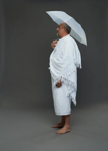 A portrait of a Saudi man in Ihram clothing holding a white umbrella while performing rituals, with various gestures and signals, full body, gray background.