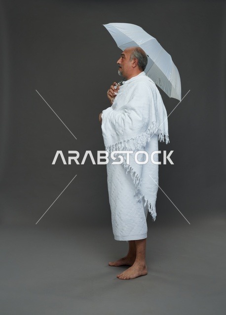 بورتريه لرجل سعودي بملابس الإحرام يحمل مظلة بيضاء أثناء أداء المناسك, ايماءات واشارات مختلفه, كامل الجسم, خلفيه رماديه
