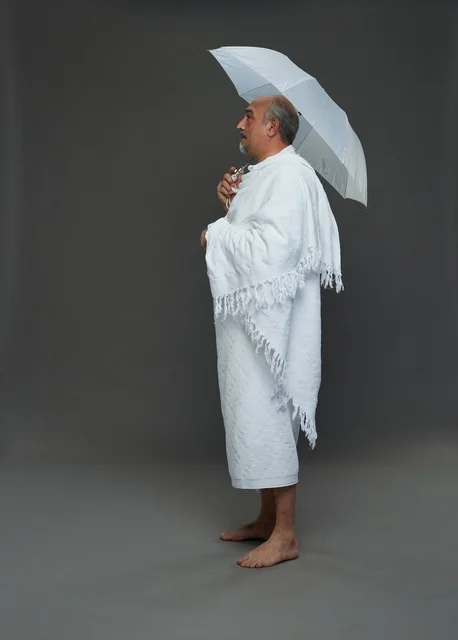 A portrait of a Saudi man in Ihram clothing holding a white umbrella while performing rituals, with various gestures and signals, full body, gray background.