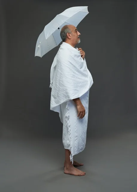 A portrait of a Saudi man in Ihram clothing holding a white umbrella while performing rituals, with various gestures and signals, full body, gray background.