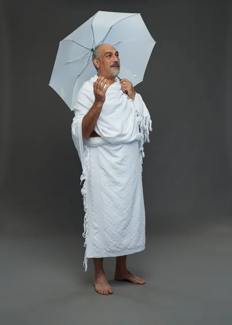 A portrait of a Saudi man in Ihram clothing holding a white umbrella while performing rituals, with various gestures and signals, full body, gray background.