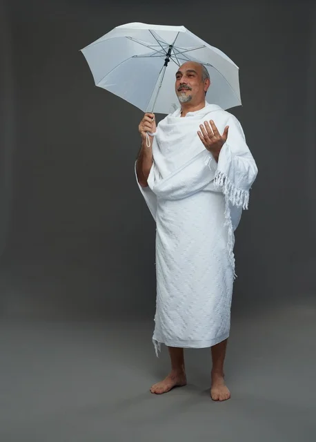 A portrait of a Saudi man in Ihram clothing holding a white umbrella while performing rituals, with various gestures and signals, full body, gray background.