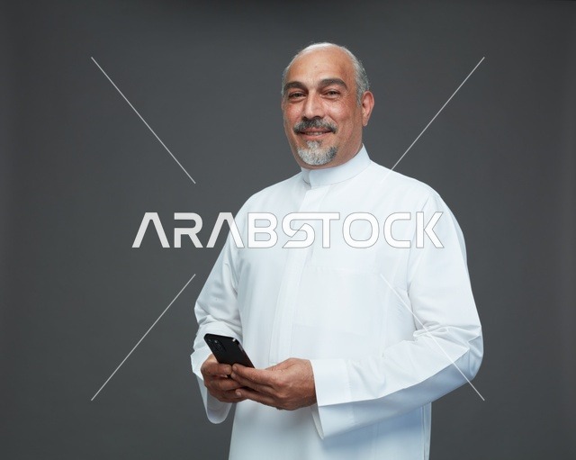 An elderly Arab Saudi Gulf man wearing a white thobe and holding a mobile phone, making various gestures and signals, full body, gray background.