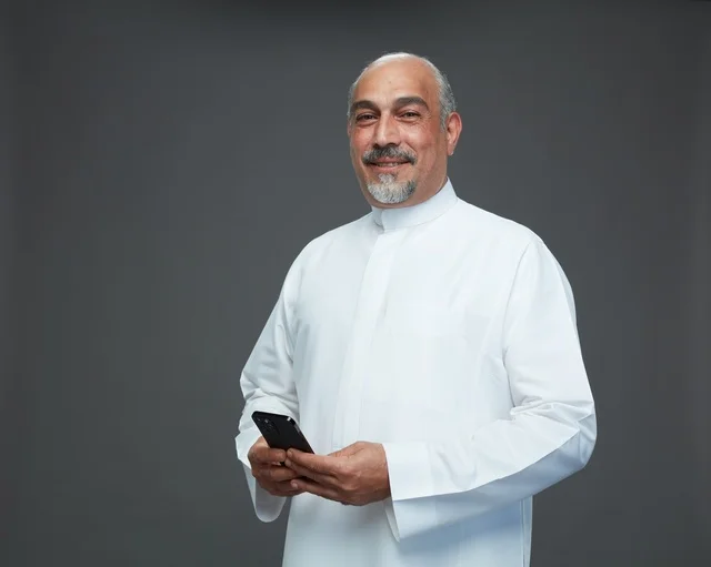 An elderly Arab Saudi Gulf man wearing a white thobe and holding a mobile phone, making various gestures and signals, full body, gray background.