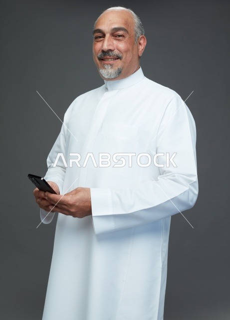رجل  عربي سعودي خليجي مسن يرتدي الزي الثوب الابيض ويمسك الهاتف المحمول,  ايماءات واشارات مختلفه , كامل الجسم, خلفيه رماديه