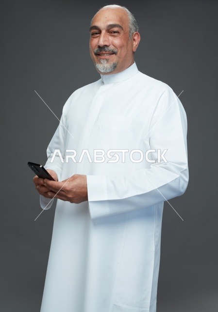 رجل  عربي سعودي خليجي مسن يرتدي الزي الثوب الابيض ويمسك الهاتف المحمول,  ايماءات واشارات مختلفه , كامل الجسم, خلفيه رماديه