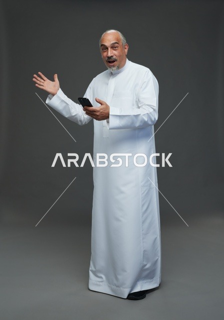 An elderly Arab Saudi Gulf man wearing a white thobe and holding a mobile phone, making various gestures and signals, full body, gray background.
