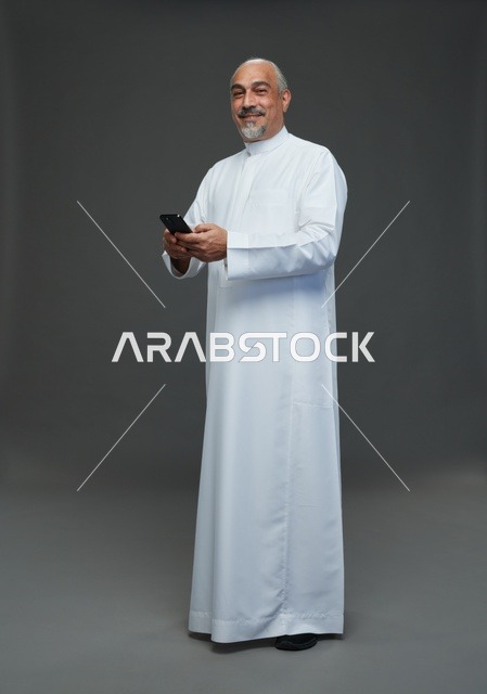 رجل  عربي سعودي خليجي مسن يرتدي الزي الثوب الابيض ويمسك الهاتف المحمول,  ايماءات واشارات مختلفه , كامل الجسم, خلفيه رماديه