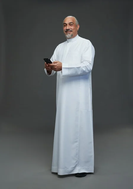 An elderly Arab Saudi Gulf man wearing a white thobe and holding a mobile phone, making various gestures and signals, full body, gray background.
