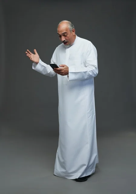 رجل  عربي سعودي خليجي مسن يرتدي الزي الثوب الابيض ويمسك الهاتف المحمول,  ايماءات واشارات مختلفه , كامل الجسم, خلفيه رماديه