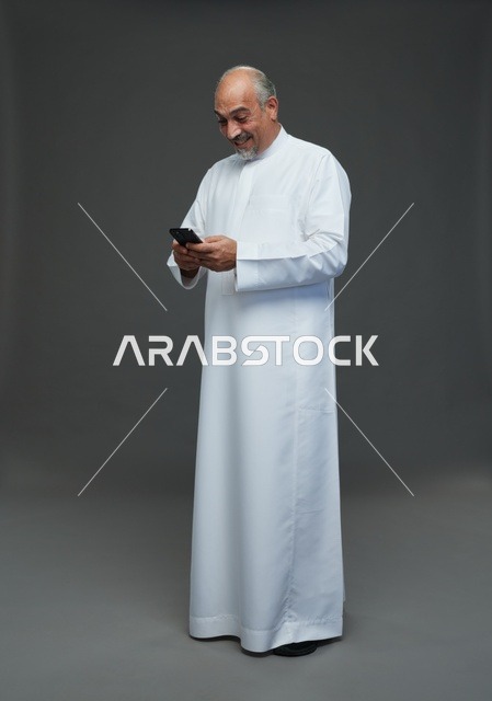 An elderly Arab Saudi Gulf man wearing a white thobe and holding a mobile phone, making various gestures and signals, full body, gray background.