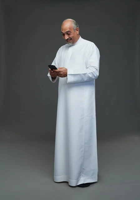 An elderly Arab Saudi Gulf man wearing a white thobe and holding a mobile phone, making various gestures and signals, full body, gray background.