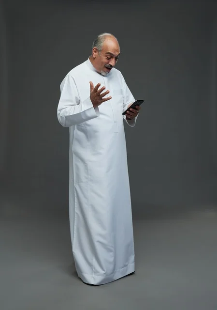An elderly Arab Saudi Gulf man wearing a white thobe and holding a mobile phone, making various gestures and signals, full body, gray background.