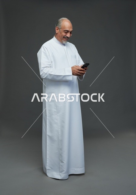 رجل  عربي سعودي خليجي مسن يرتدي الزي الثوب الابيض ويمسك الهاتف المحمول,  ايماءات واشارات مختلفه , كامل الجسم, خلفيه رماديه
