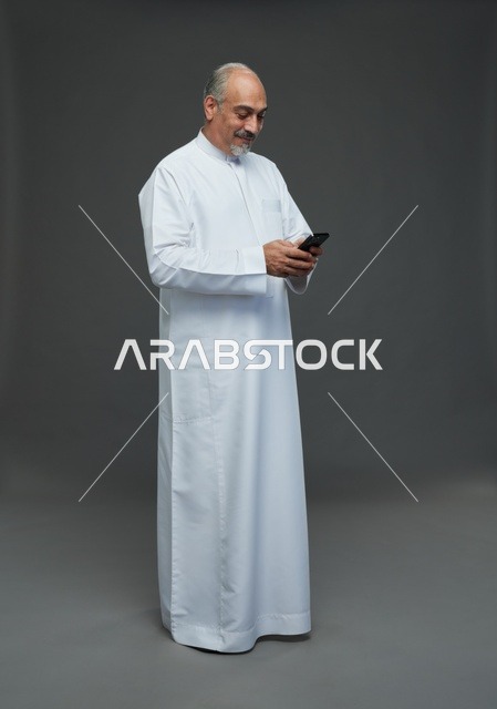رجل  عربي سعودي خليجي مسن يرتدي الزي الثوب الابيض ويمسك الهاتف المحمول,  ايماءات واشارات مختلفه , كامل الجسم, خلفيه رماديه