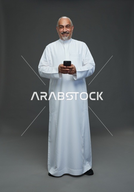 An elderly Arab Saudi Gulf man wearing a white thobe and holding a mobile phone, making various gestures and signals, full body, gray background.