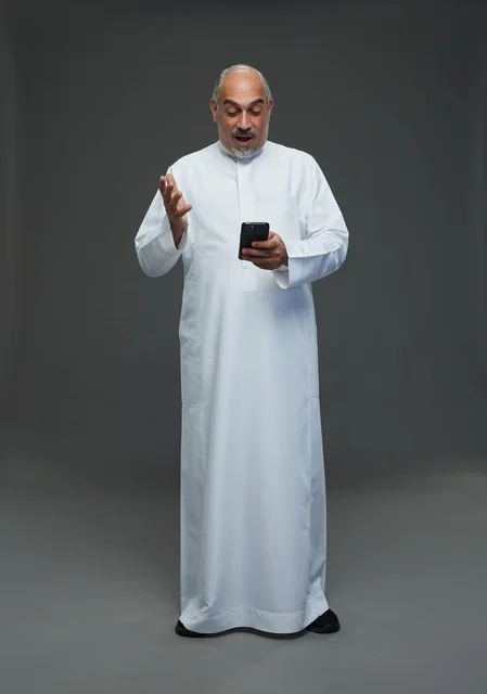 An elderly Arab Saudi Gulf man wearing a white thobe and holding a mobile phone, making various gestures and signals, full body, gray background.
