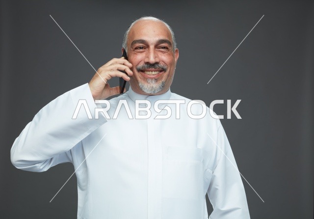 رجل  عربي سعودي خليجي مسن يرتدي الزي الثوب الابيض ويمسك الهاتف المحمول, اجراء مكالمه هاتفيه,  ايماءات واشارات مختلفه , كامل الجسم, خلفيه رماديه