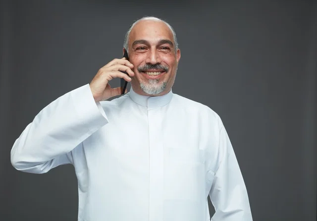 رجل  عربي سعودي خليجي مسن يرتدي الزي الثوب الابيض ويمسك الهاتف المحمول, اجراء مكالمه هاتفيه,  ايماءات واشارات مختلفه , كامل الجسم, خلفيه رماديه