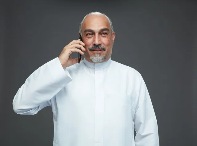 An elderly Arab Saudi Gulf man wearing a white thobe is holding a mobile phone, making a phone call, with various gestures and signals, full body, gray background.