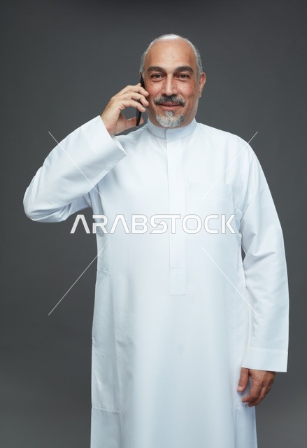 An elderly Arab Saudi Gulf man wearing a white thobe is holding a mobile phone, making a phone call, with various gestures and signals, full body, gray background.