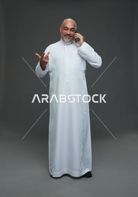 رجل  عربي سعودي خليجي مسن يرتدي الزي الثوب الابيض ويمسك الهاتف المحمول, اجراء مكالمه هاتفيه,  ايماءات واشارات مختلفه , كامل الجسم, خلفيه رماديه