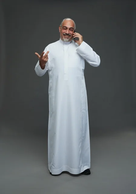 An elderly Arab Saudi Gulf man wearing a white thobe is holding a mobile phone, making a phone call, with various gestures and signals, full body, gray background.