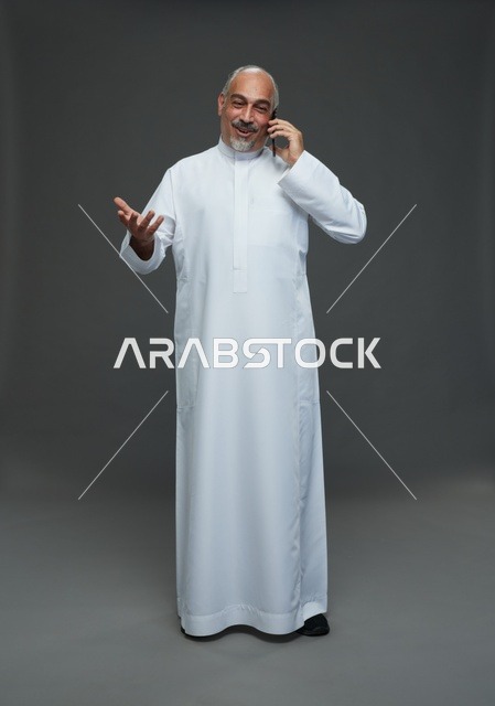 An elderly Arab Saudi Gulf man wearing a white thobe is holding a mobile phone, making a phone call, with various gestures and signals, full body, gray background.